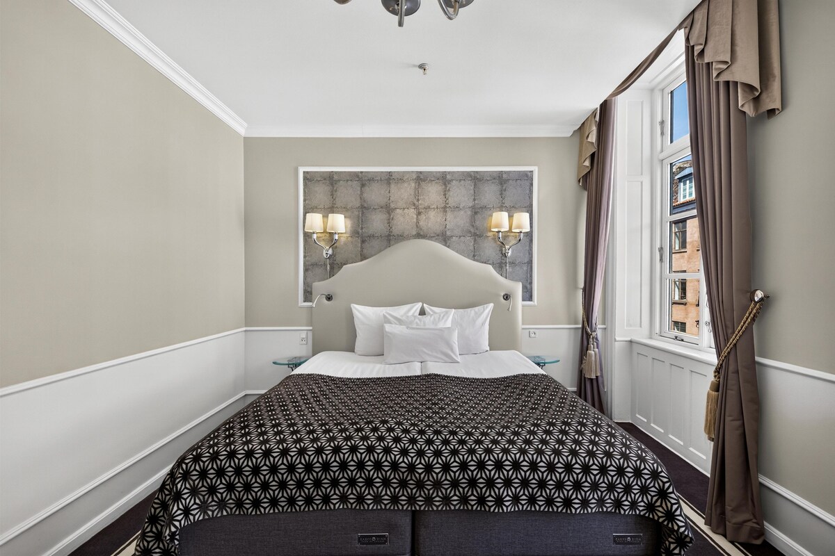 A hotel room with a bed, two bedside lamps, a window with curtains, and a view of buildings outside.
