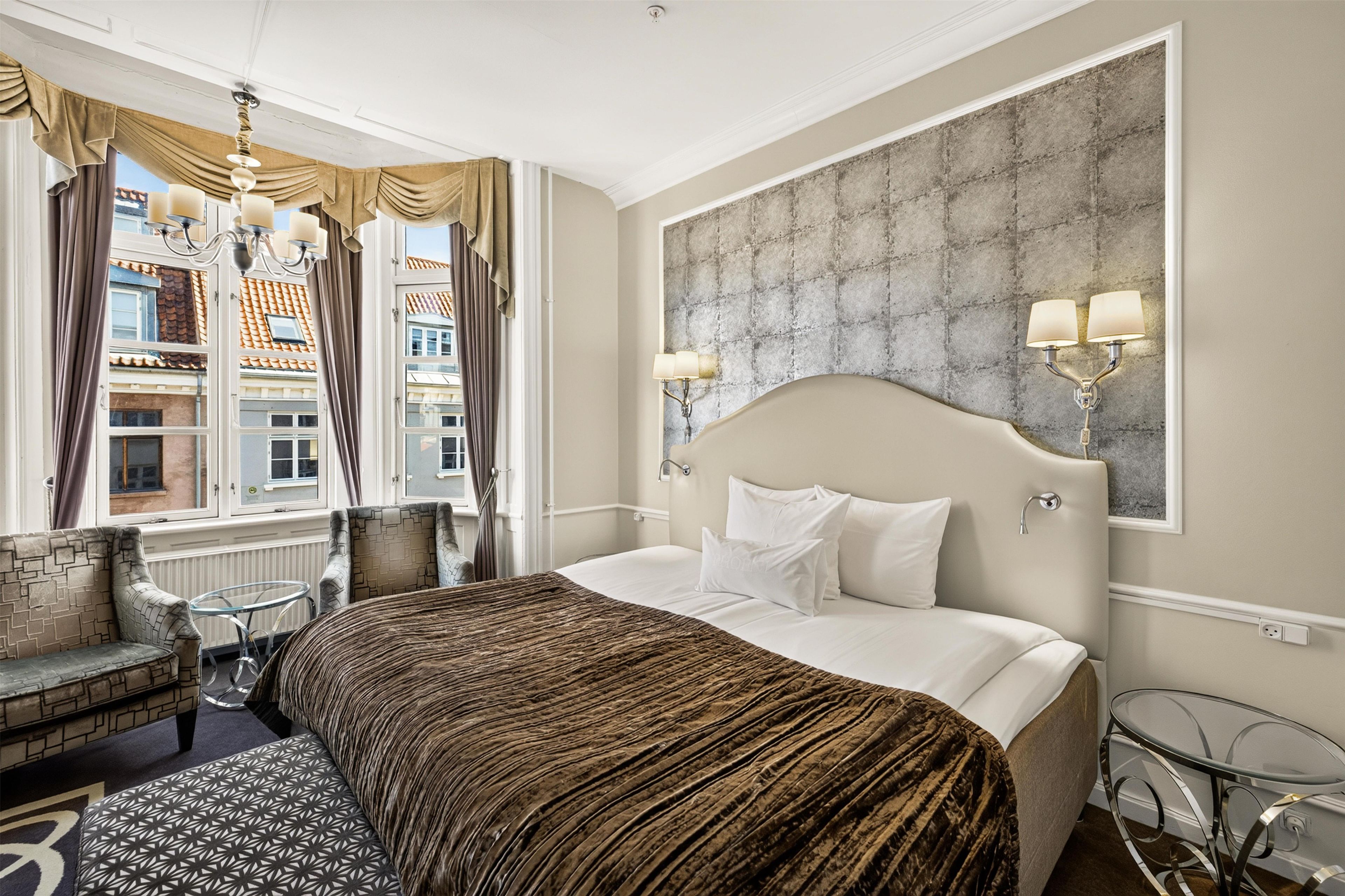 A hotel room with a large bed, a chaise lounge, a small table, and a view of buildings through the window.