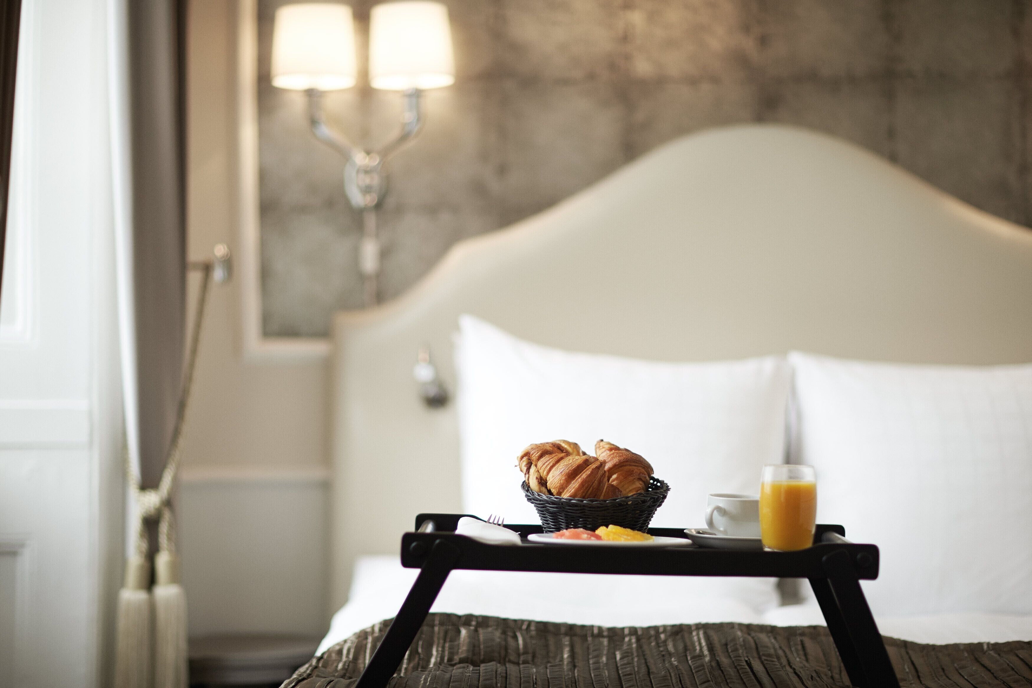 Un lit bien fait, avec du linge de lit blanc, une table de chevet sur laquelle se trouve un plateau avec des croissants et du jus d’orange, et une lampe fixée au mur.