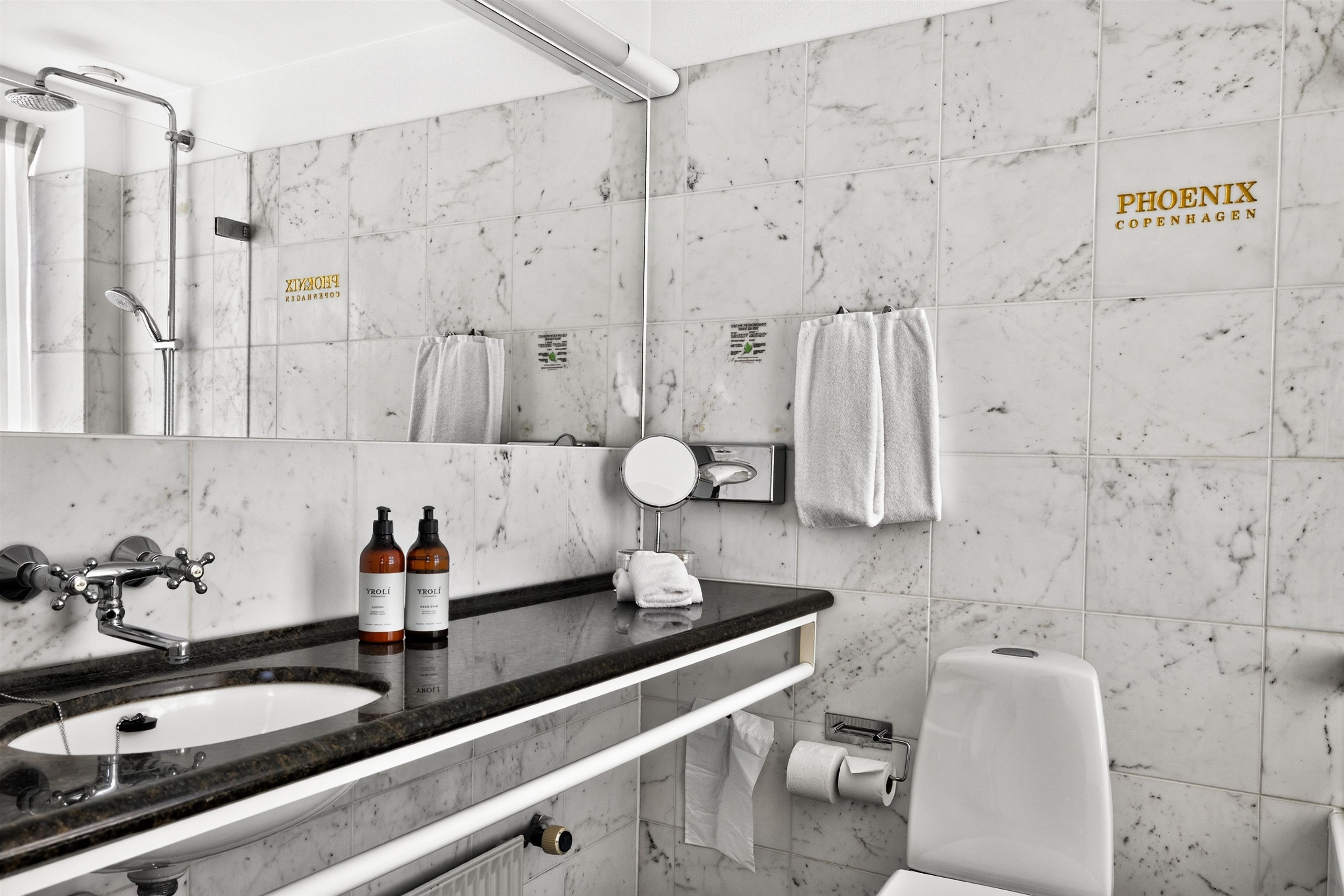 A bathroom with a marble-tiled wall, a sink with a faucet, two bottles of shampoo, a towel rack with towels, and a toilet paper holder.