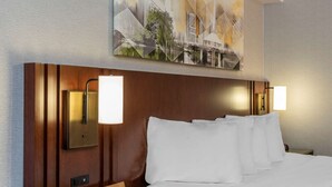 Habitación de hotel con cabecera de madera, dos lámparas de noche, cama con ropa de cama blanca y un cuadro sobre la cama.