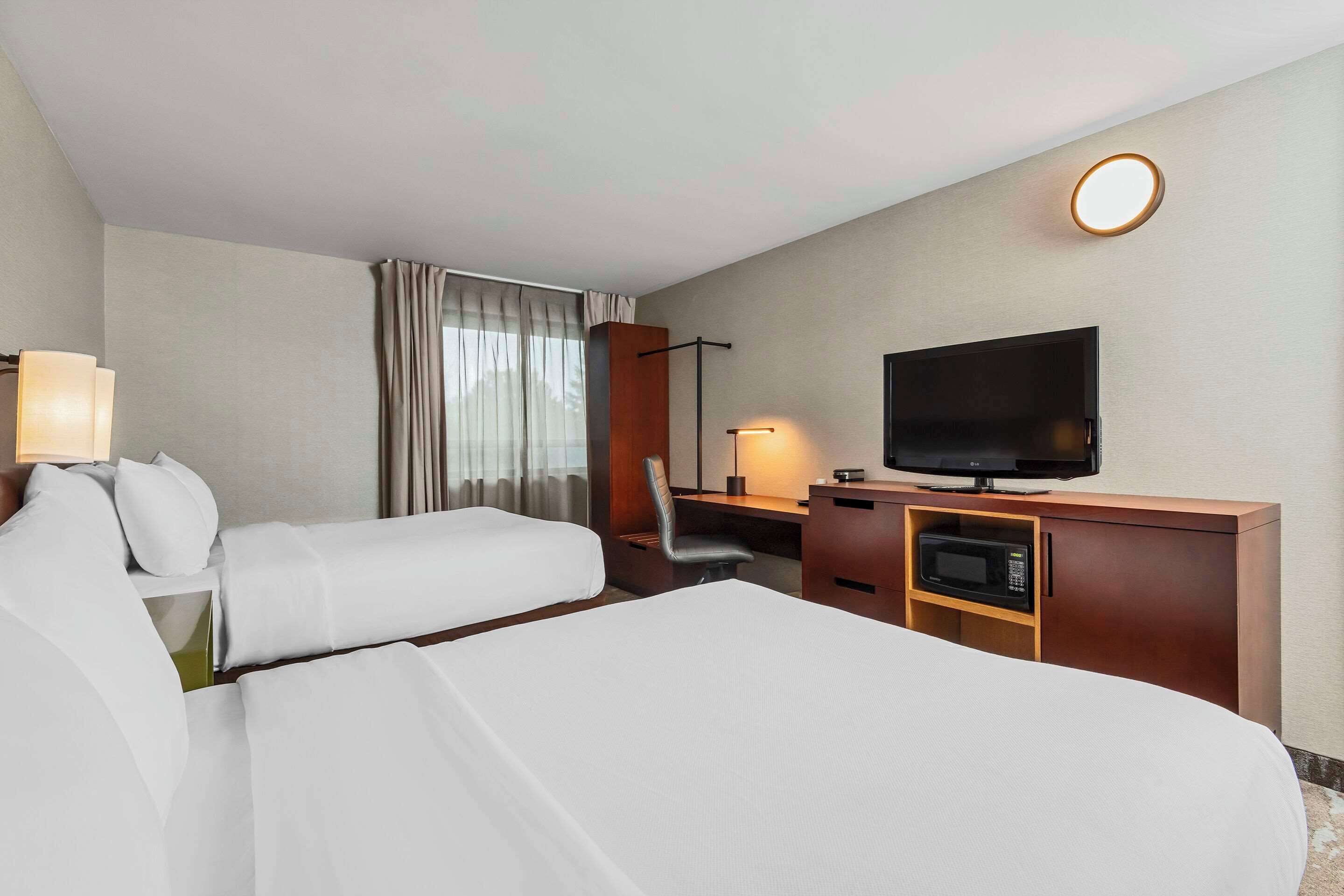 A hotel room with two beds, a desk with a television, a desk lamp, a chair, and a microwave.