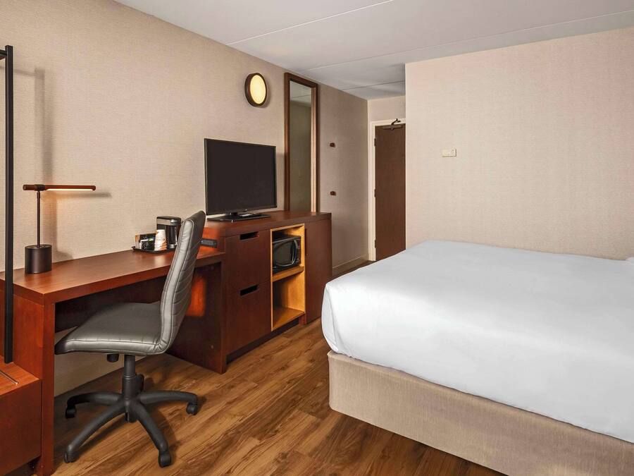 Quarto de hotel com escrivaninha de madeira, uma cadeira, cama, televisão e espelho.