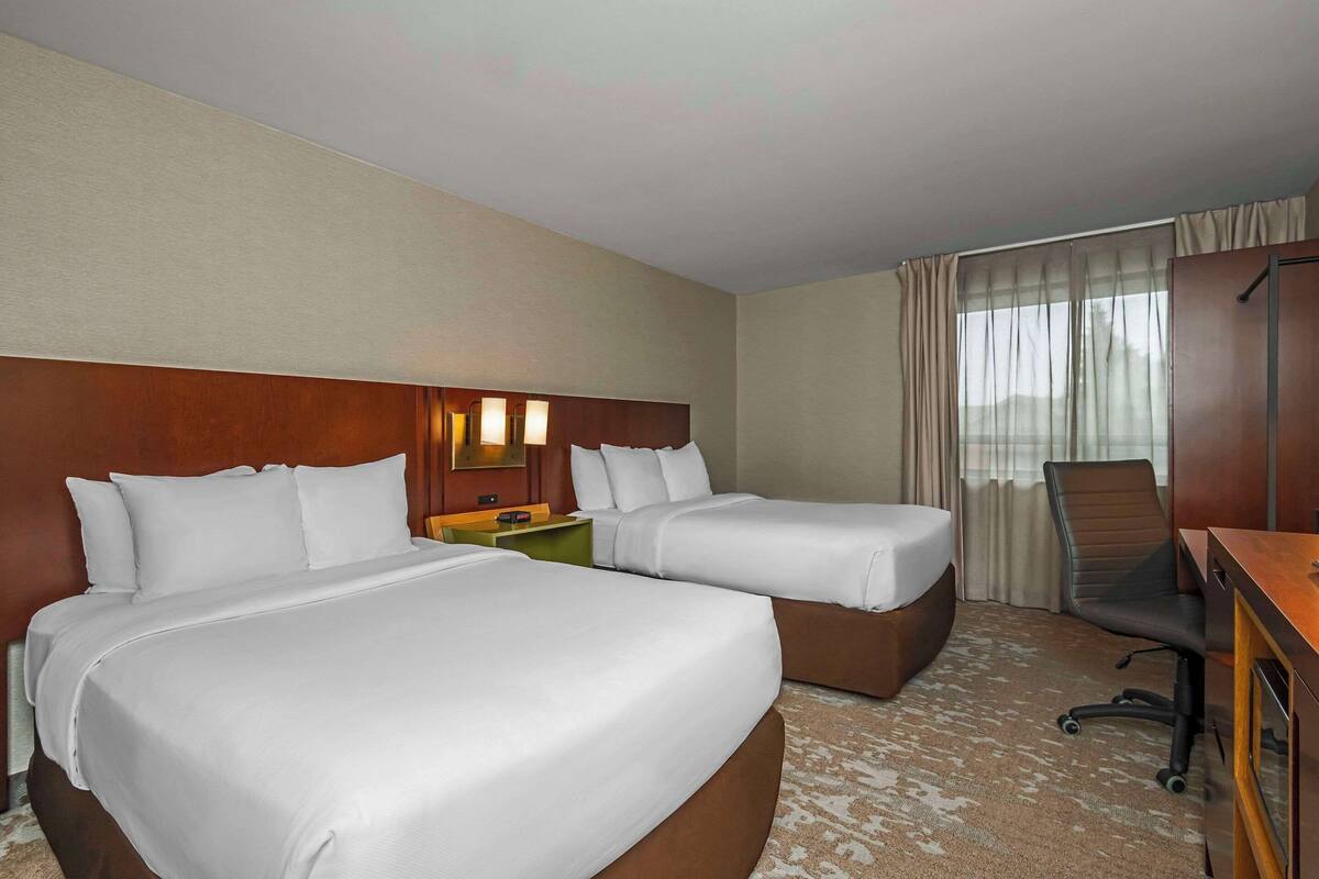 Una habitación de hotel con dos camas, un escritorio, una silla y un ventanal con cortinas.