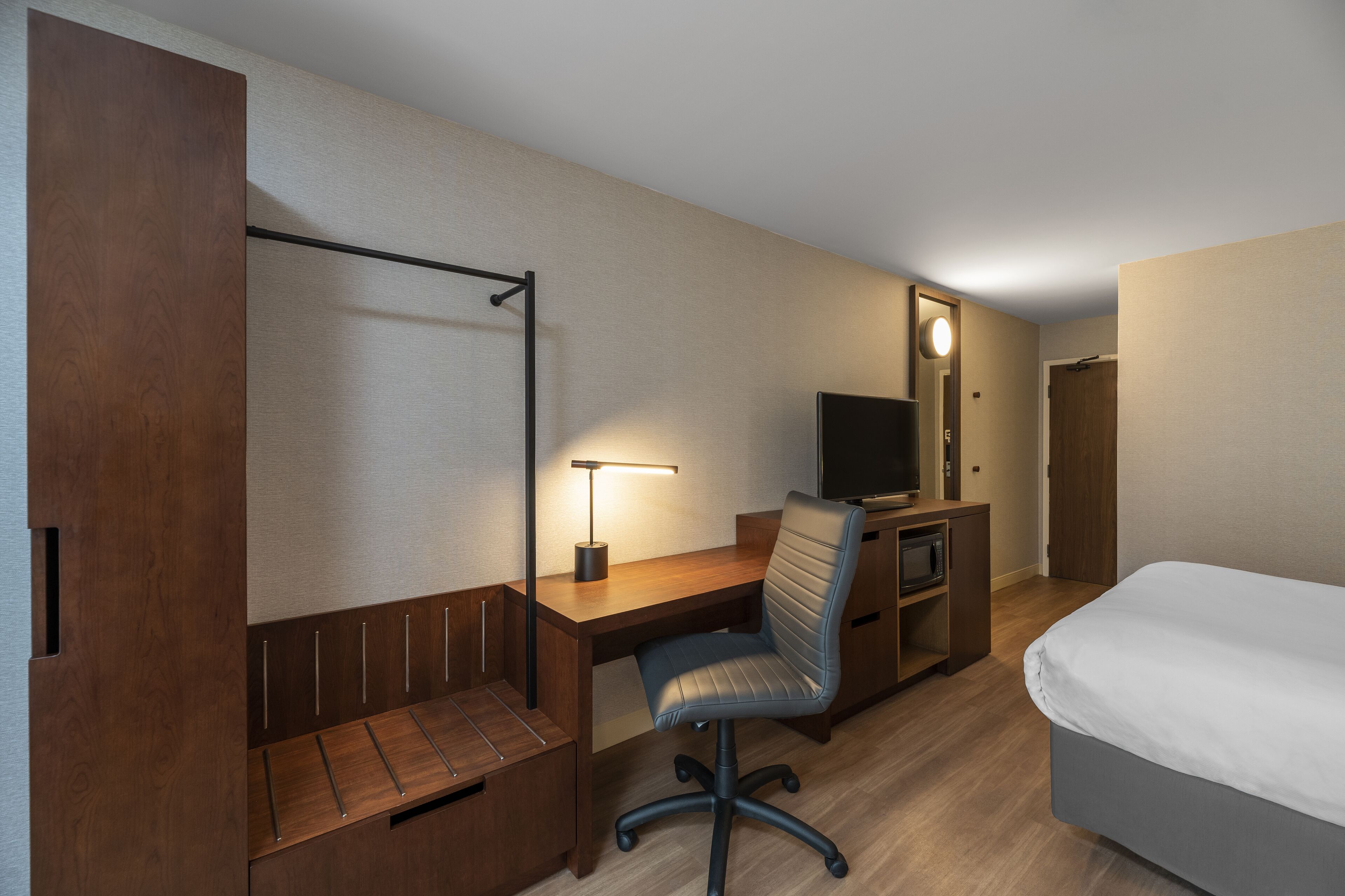 A hotel room with a wooden desk, a chair, a television, a bed, and a wardrobe.