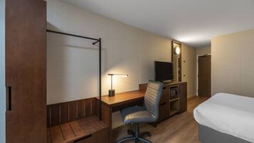 A hotel room with a wooden desk, a chair, a television, a bed, and a wardrobe.