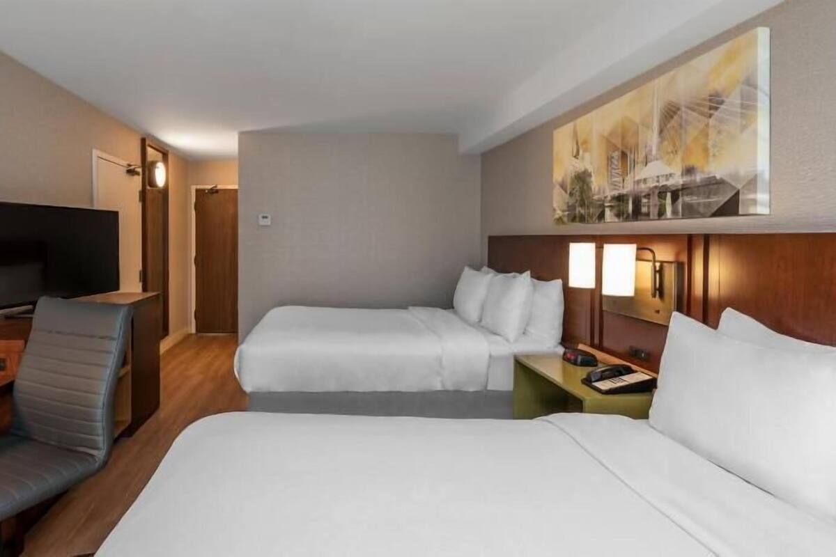Habitación de hotel con dos camas, un escritorio con silla, un televisor y un cuadro en la pared.
