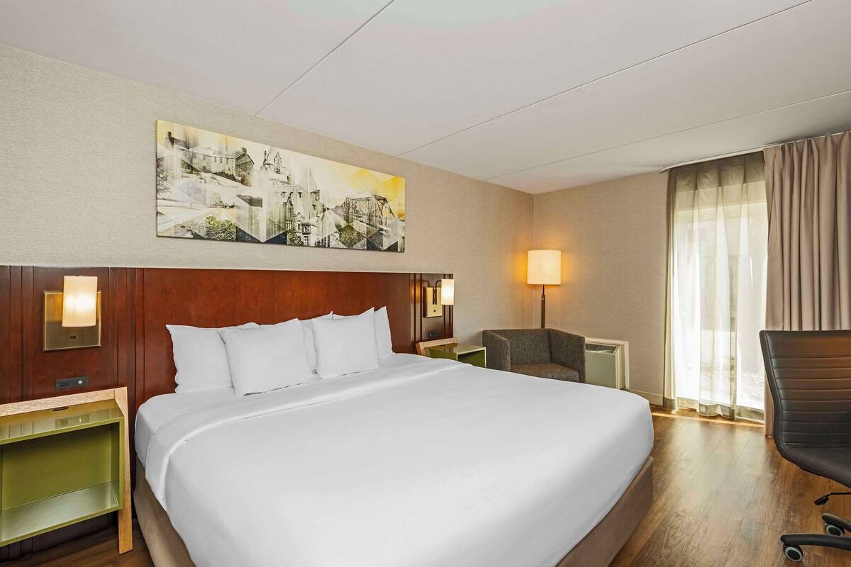 Habitación de hotel con una cama grande, cabecera con un cuadro, dos mesitas de noche con lámparas, una silla y una ventana con cortinas.