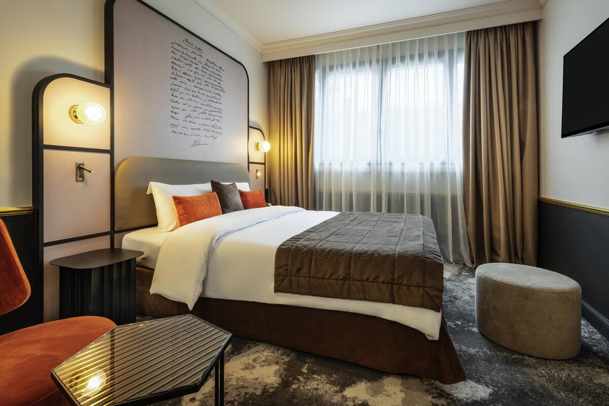 A hotel room with a large bed, a bedside table, a chair, a small ottoman, a TV mounted on the wall, and a window with curtains.