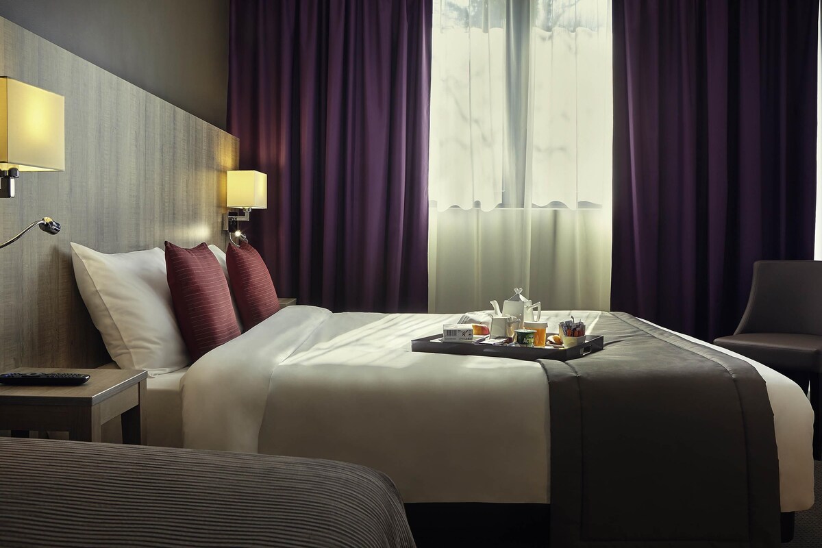 A hotel room with a bed, bedside lamps, a tray with breakfast items, and a view through the window.