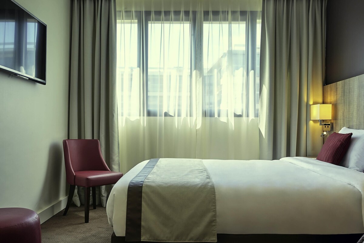 Ett hotellrum med en säng, en stol, en tv och ett fönster med gardiner.