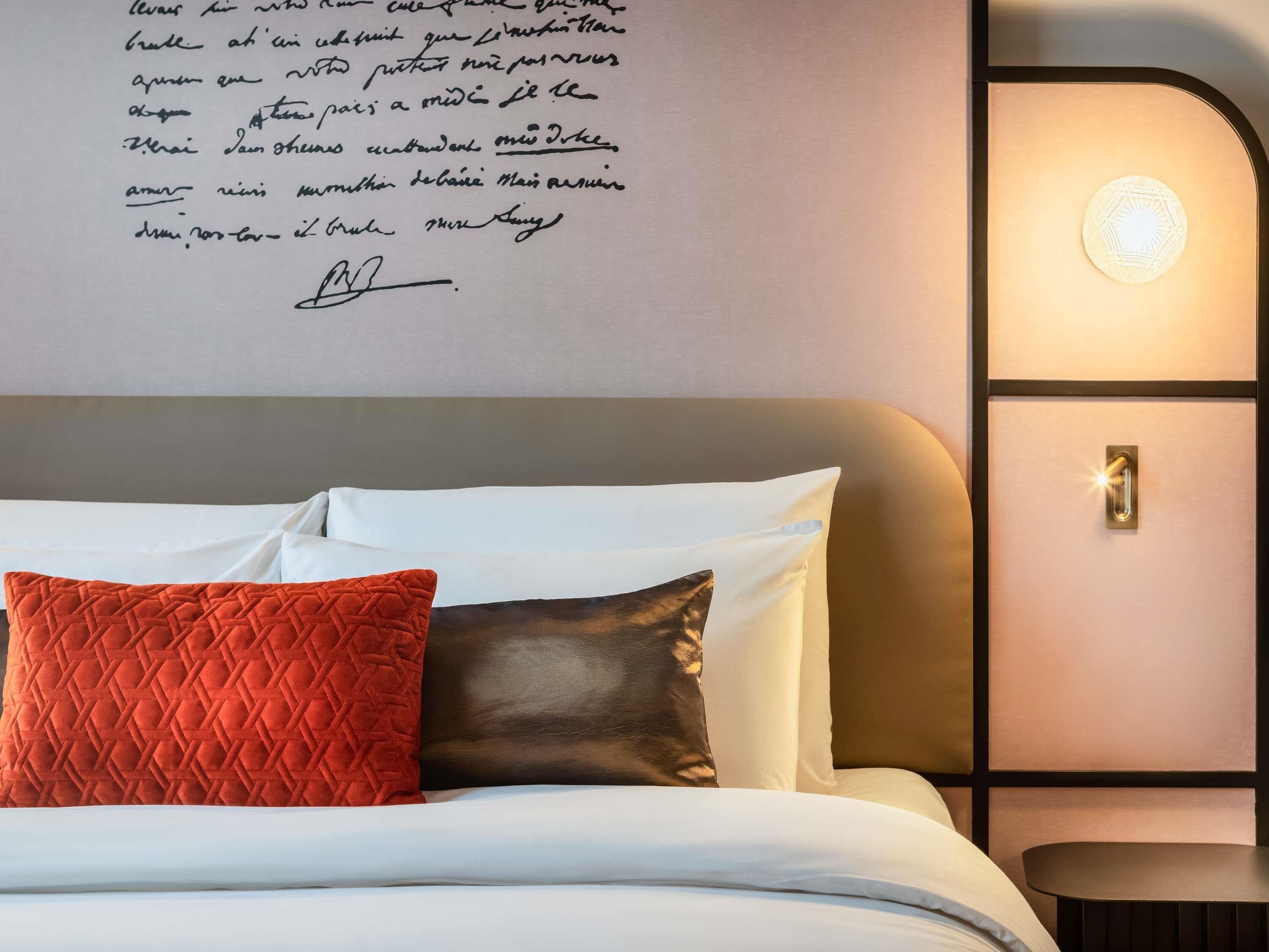 A neatly made bed with white and brown pillows, a red quilted pillow, and a wall with a light fixture and a door.