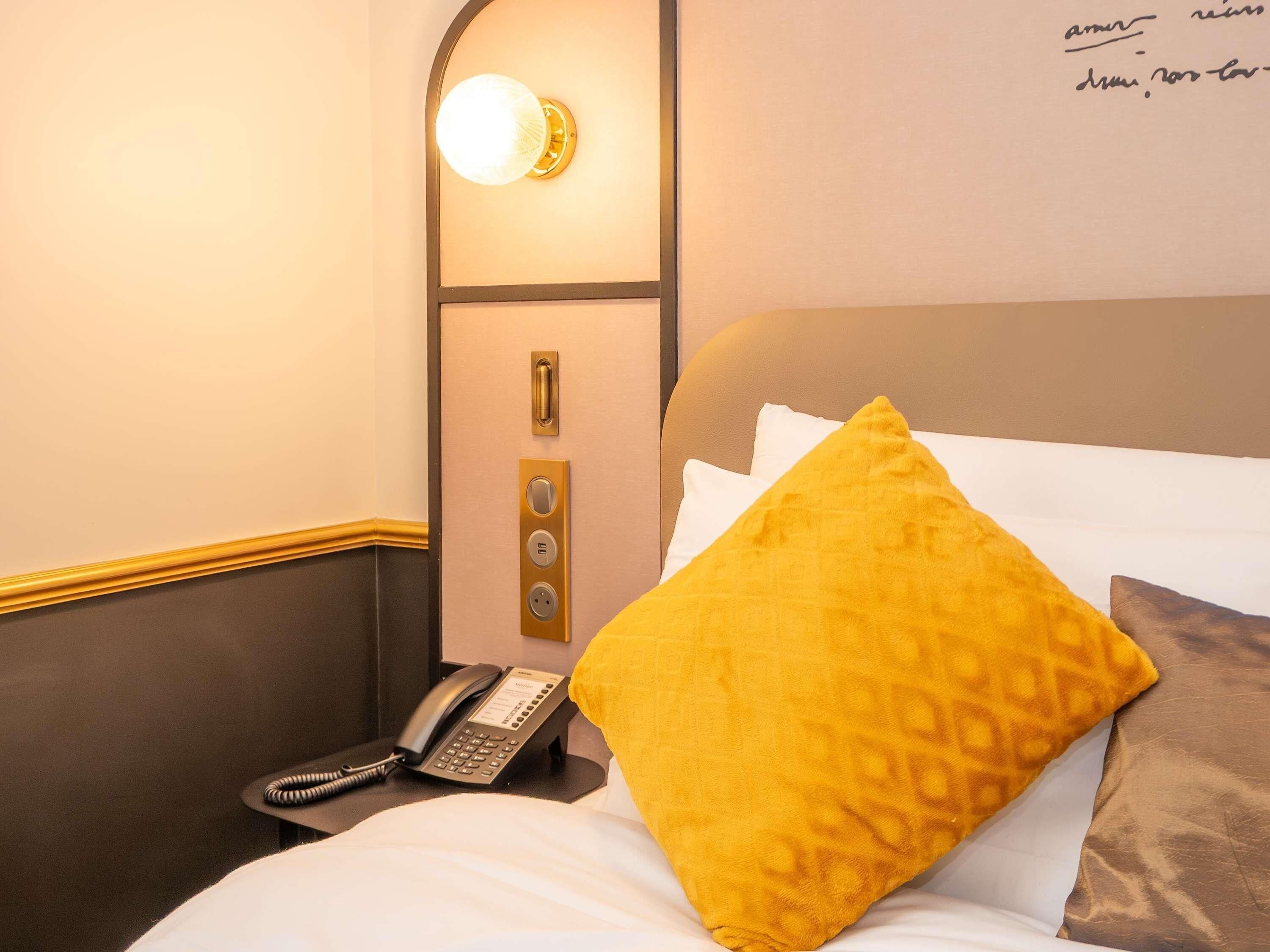 A hotel room with a bed, a bedside telephone, a wall-mounted light fixture, and a door with a handle and lock.