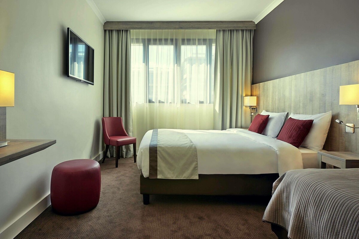 A hotel room with a bed, a chair, a red ottoman, a TV, and a window with curtains.
