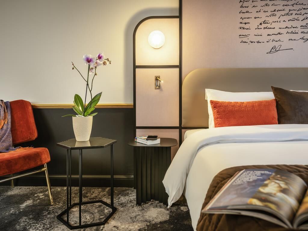 A hotel room with a bed, a small table with a plant, a chair, and a bedside table with a lamp.