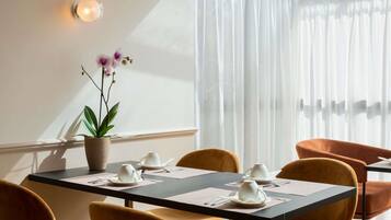 Un coin repas avec une table dressée pour deux personnes, des chaises, une plante en pot et une fenêtre ornée de rideaux légers.