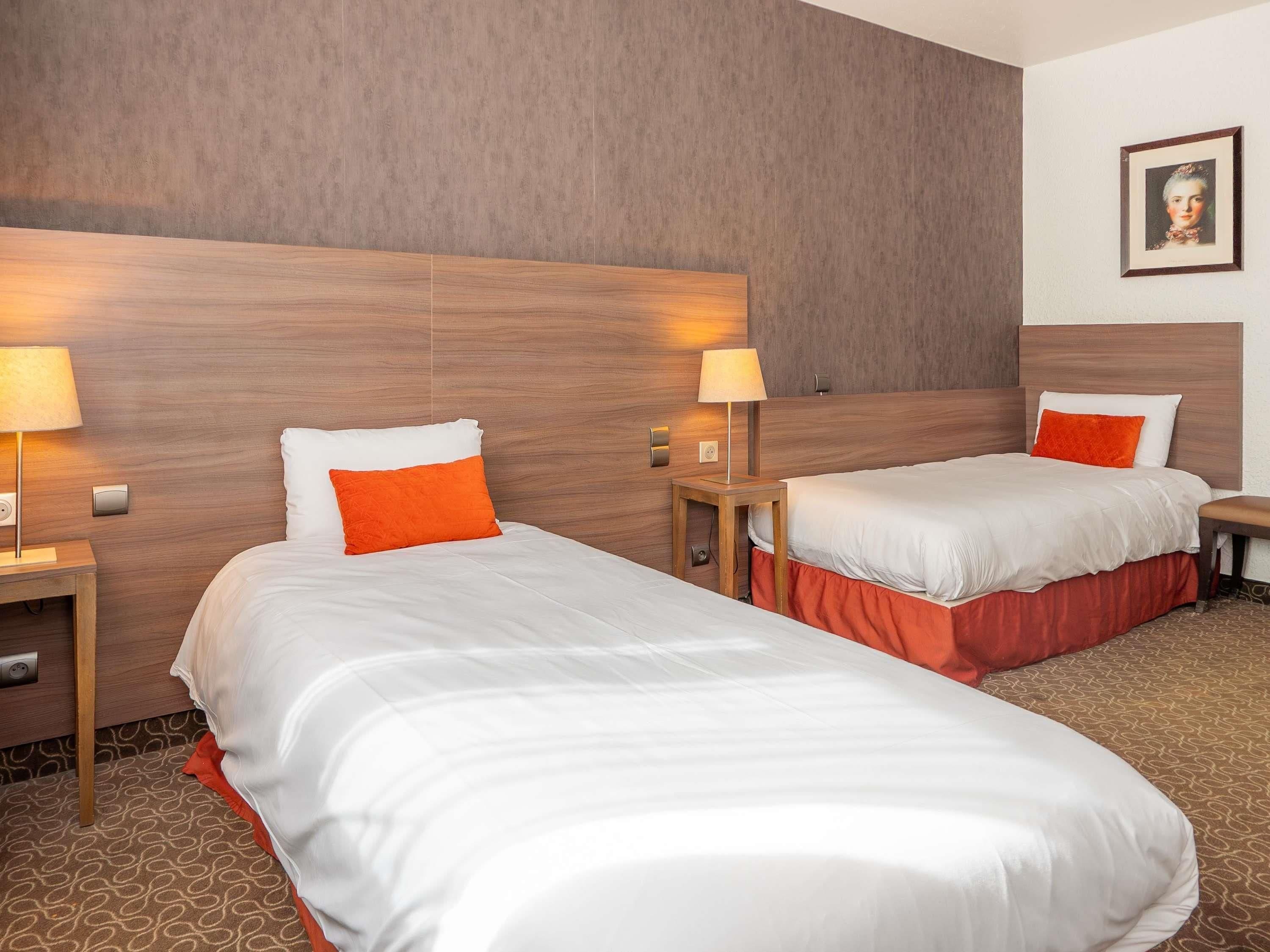 Habitación de hotel con dos camas, cada una con ropa blanca y almohadas anaranjadas, cabeceras de madera y lámparas de noche.
