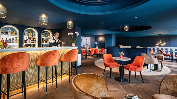 A bar area with a bartender, red and brown chairs, and a bar counter with bottles. - Mercure Versailles Paris Ouest (Le Chesnay-Rocquencourt)