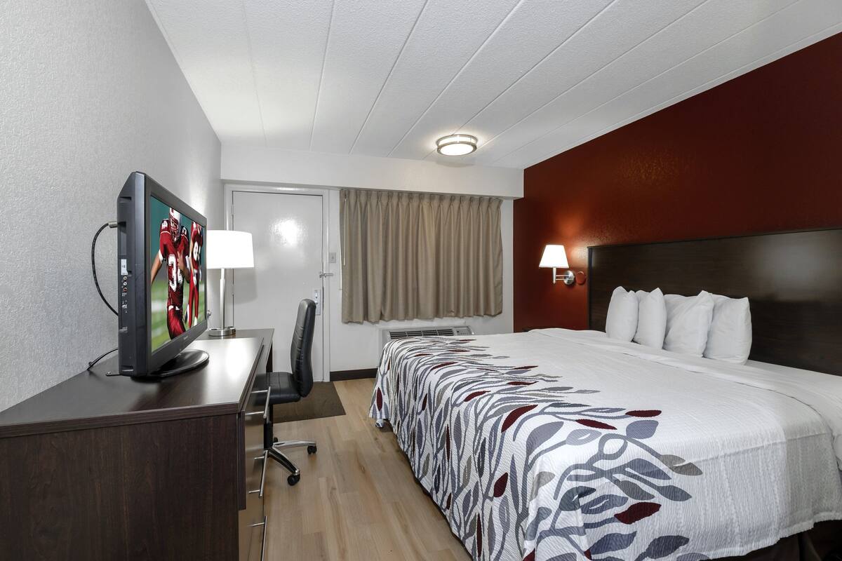 A hotel room with a bed, a desk with a TV, a chair, a lamp, and a window with curtains.
