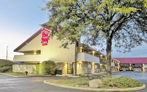 Un motel avec un panneau rouge sur le toit, un parking et un grand arbre à l’avant.