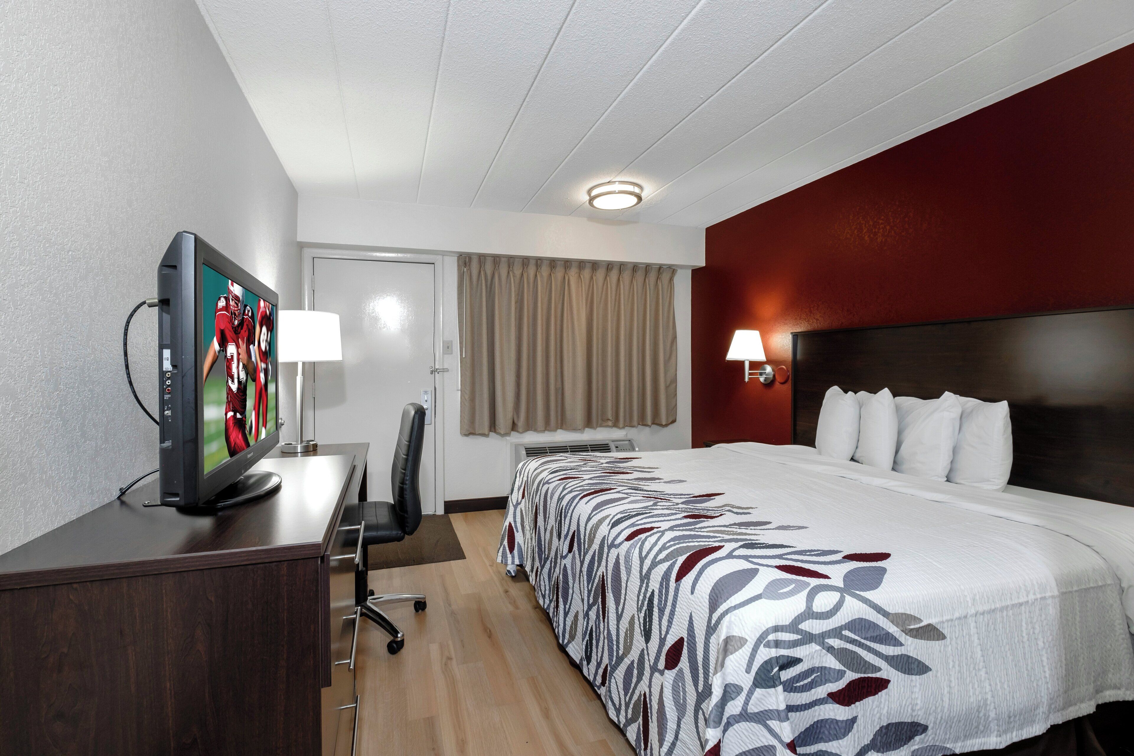 Una habitación de hotel con una cama, un escritorio con televisor, una silla, una lámpara y un ventanal con cortinas.