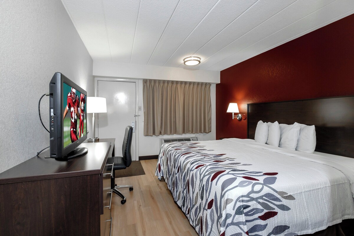 Una habitación de hotel con una cama, un escritorio con televisor, una silla, una lámpara y un ventanal con cortinas.