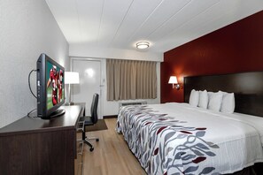 Una habitación de hotel con una cama, un escritorio con televisor, una silla, una lámpara y un ventanal con cortinas.