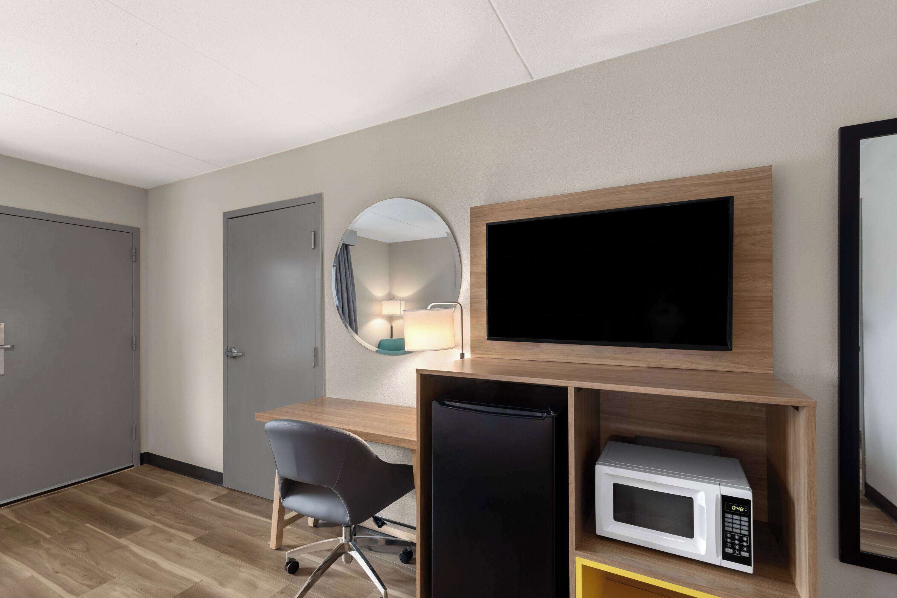 A hotel room with a wooden desk, a black refrigerator, a white microwave, a large flat-screen TV, a round mirror, and a chair.