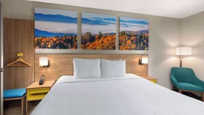 Ein Hotelzimmer mit einem großen Bett, einem hölzernen Kopfteil, einem Schreibtisch mit Lampe, einem Sessel und einem Wandbild einer Waldlandschaft.