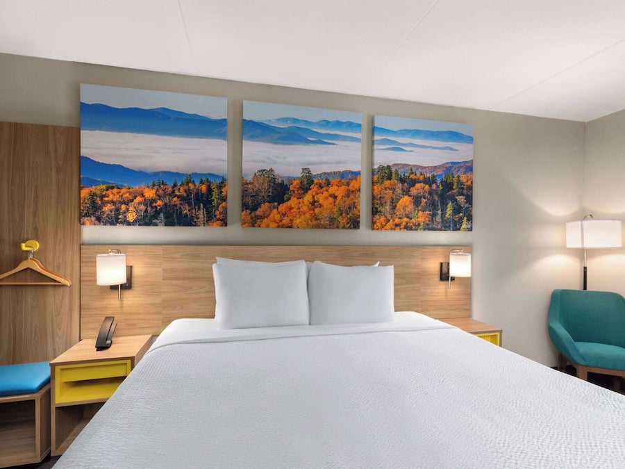 Habitación de hotel con una cama grande, cabecera de madera, escritorio con lámpara, una silla y un cuadro colgado en la pared de un paisaje forestal.