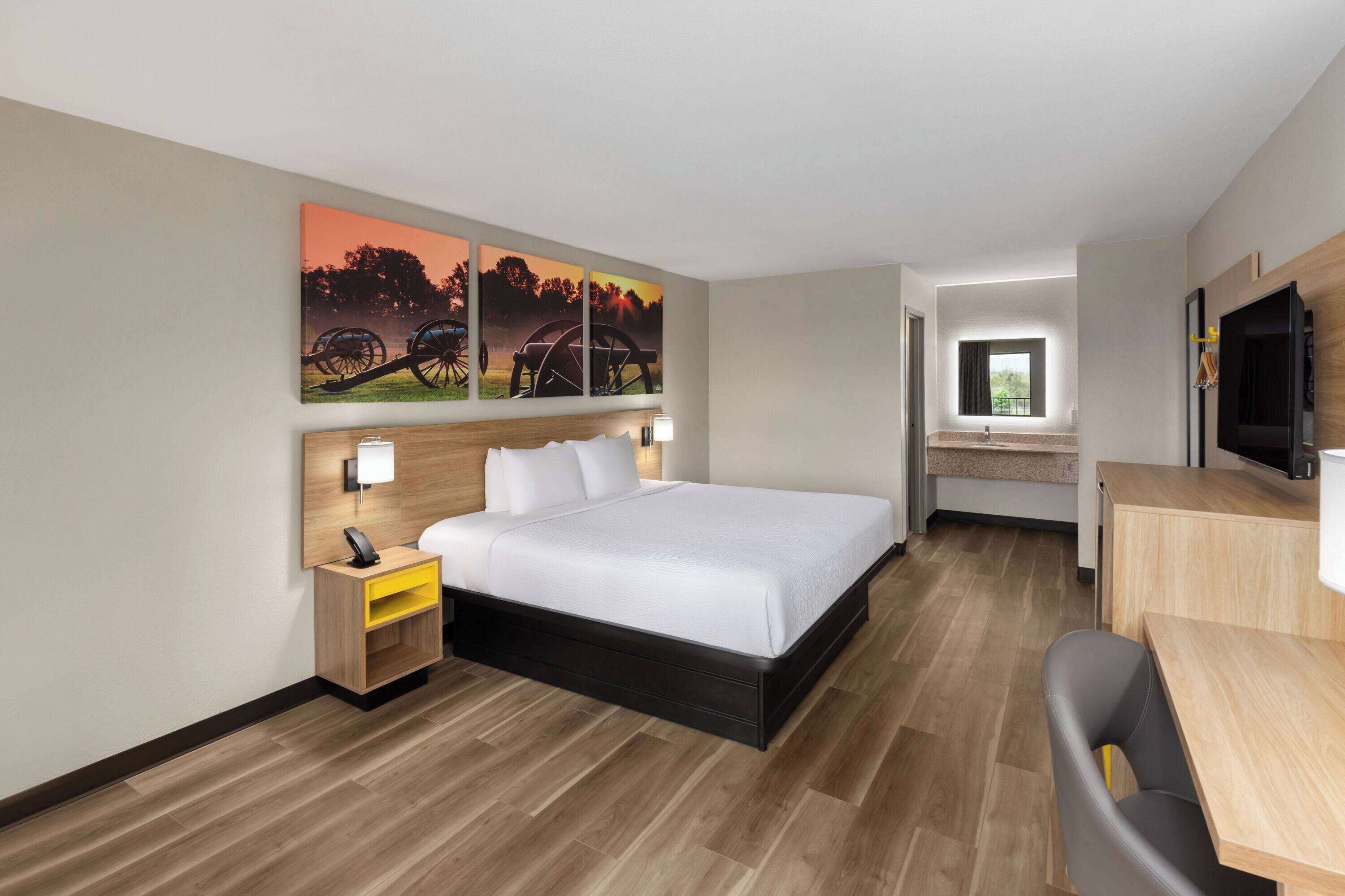 Quarto de hotel com cama, mesa de cabeceira, escrivaninha, cadeira e televisão.
