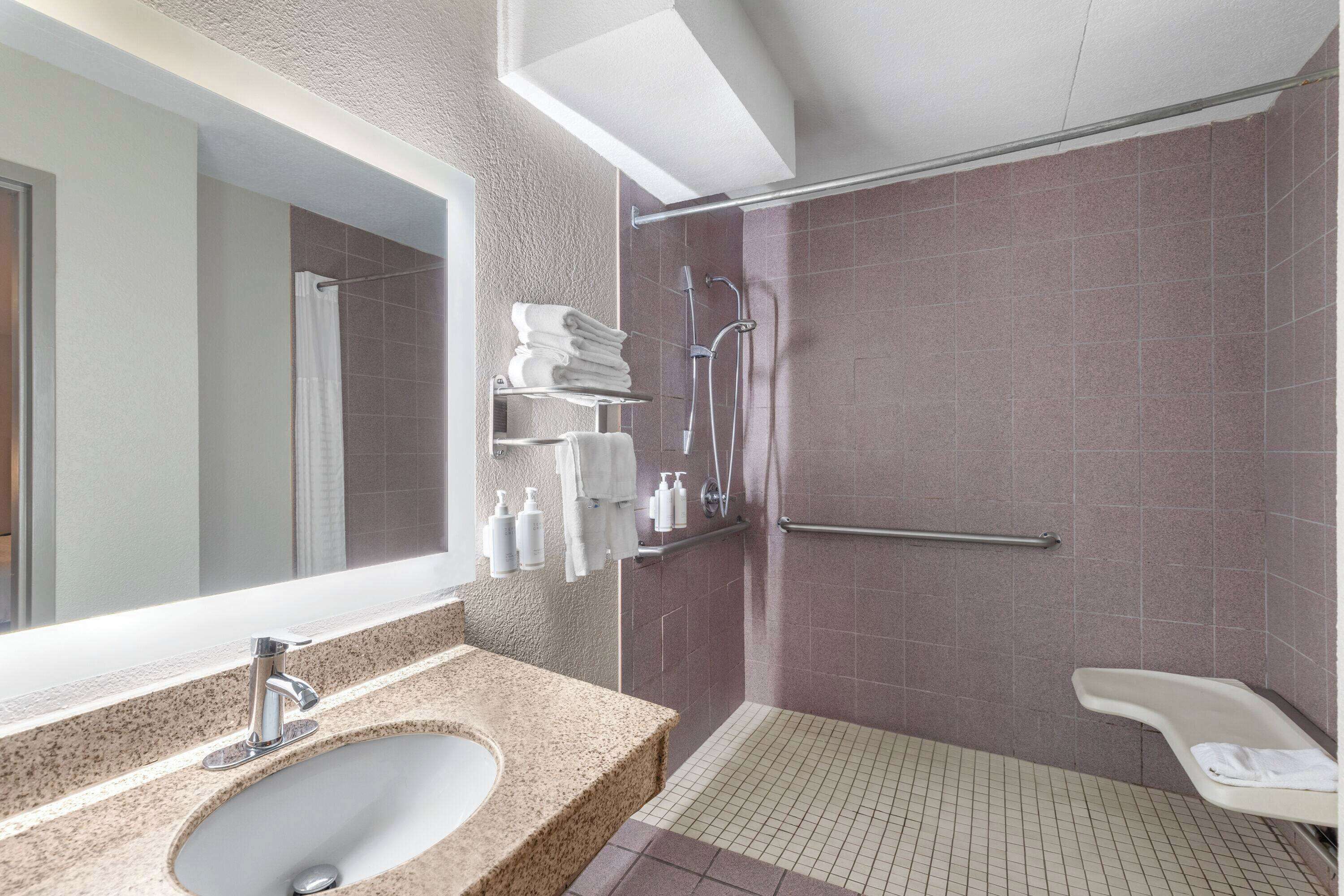 A modern bathroom with a granite sink, a large mirror, a shower with a grab bar, and a small white bench.