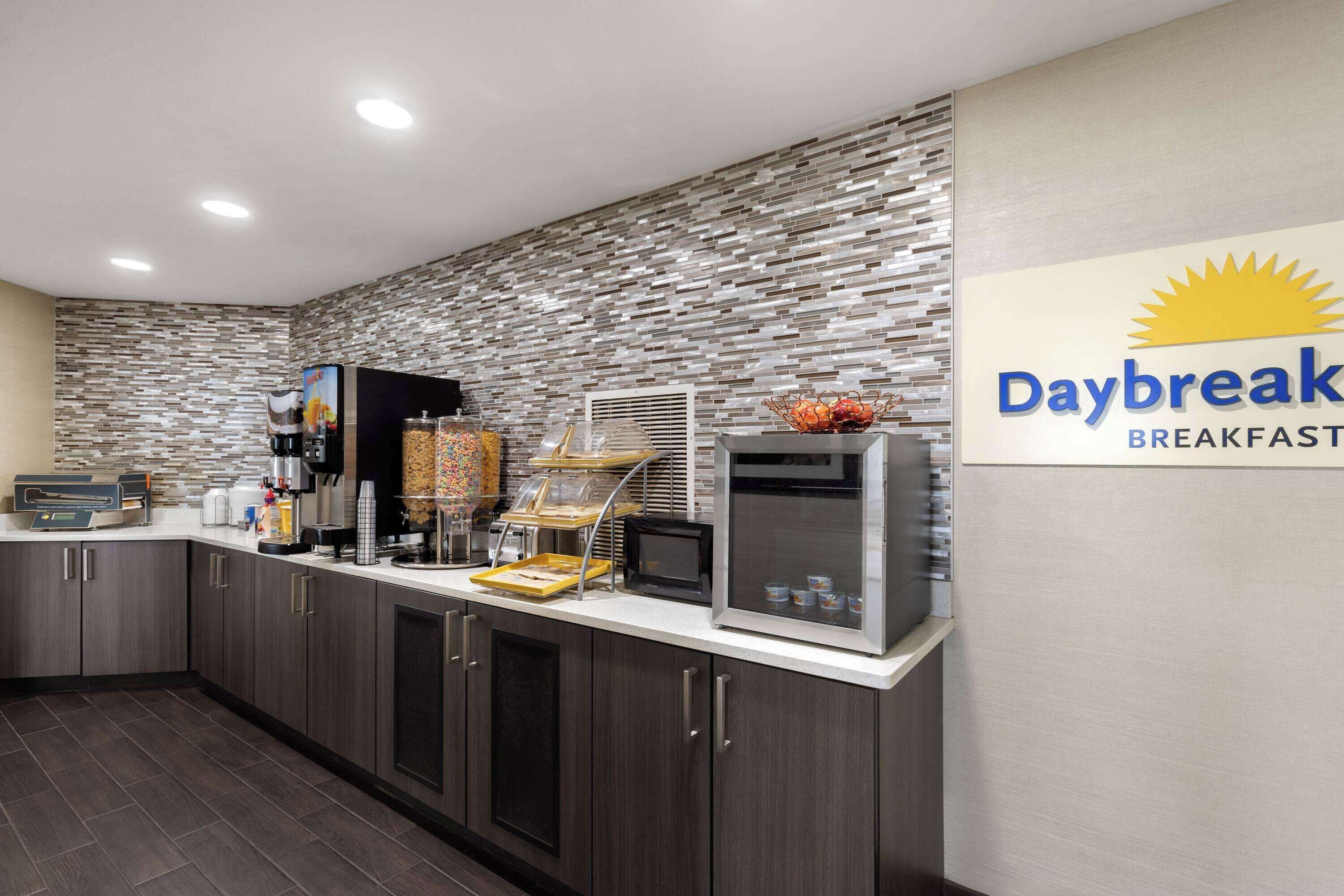 Un área de desayuno con un letrero Daybreak, una cafetera, una presentación de frutas y una barra de cereales.