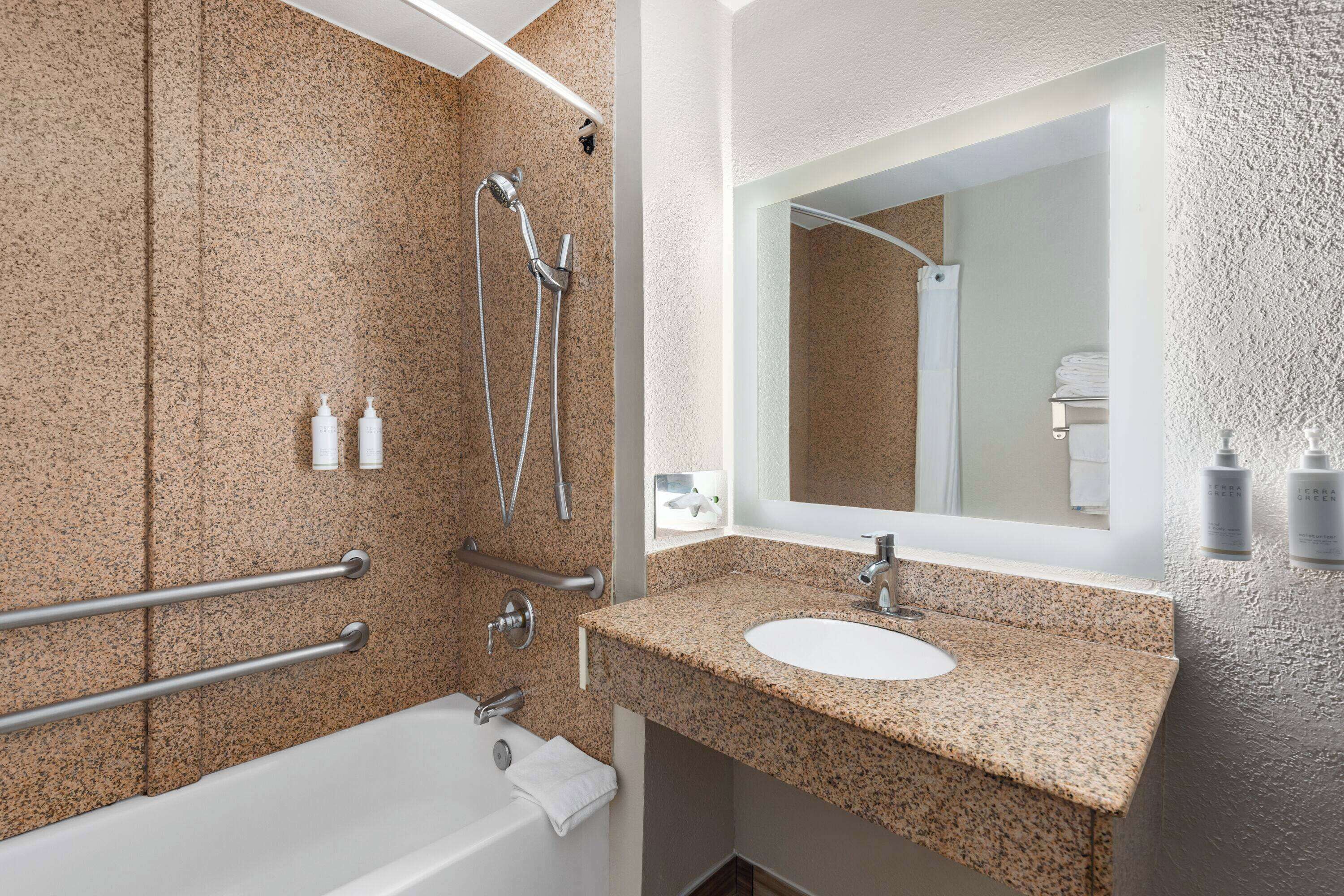 Une salle de bain moderne avec un lavabo sur un plan de travail en granit, un grand miroir et un espace douche équipé de barres d’appui.