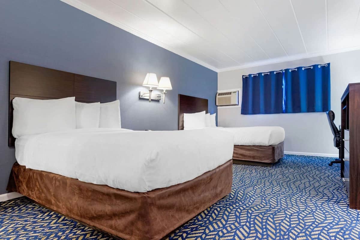 A hotel room with a large bed, a smaller bed, a desk, a chair, a lamp, and a window with curtains.