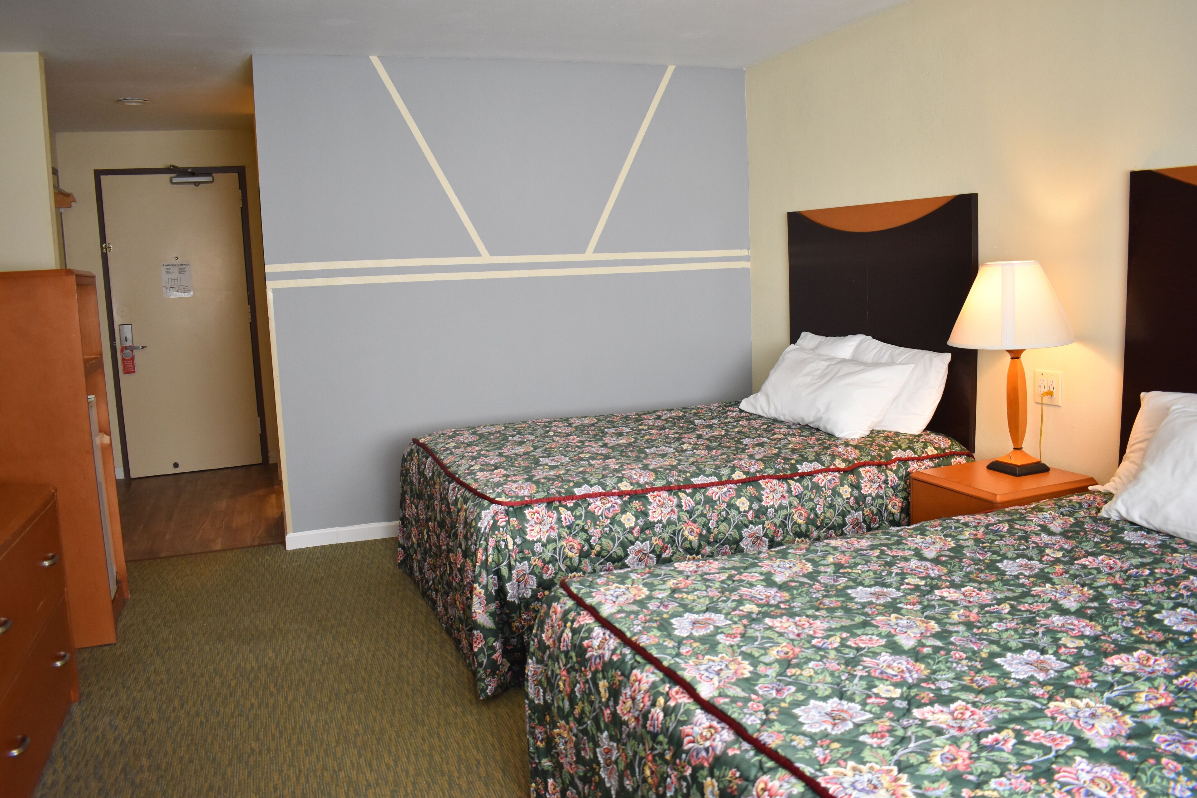 A hotel room with two beds, a dresser, a door, and a nightstand with a lamp.