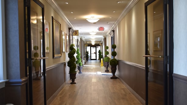 A hotel hallway with wooden flooring, framed artwork on the walls, and potted plants. - East Norwich Inn (East Norwich)