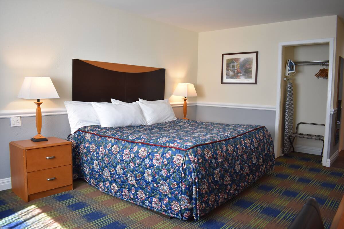 A neatly made bed with a floral bedspread, two bedside tables with lamps, a framed picture on the wall, and a door with a coat rack.