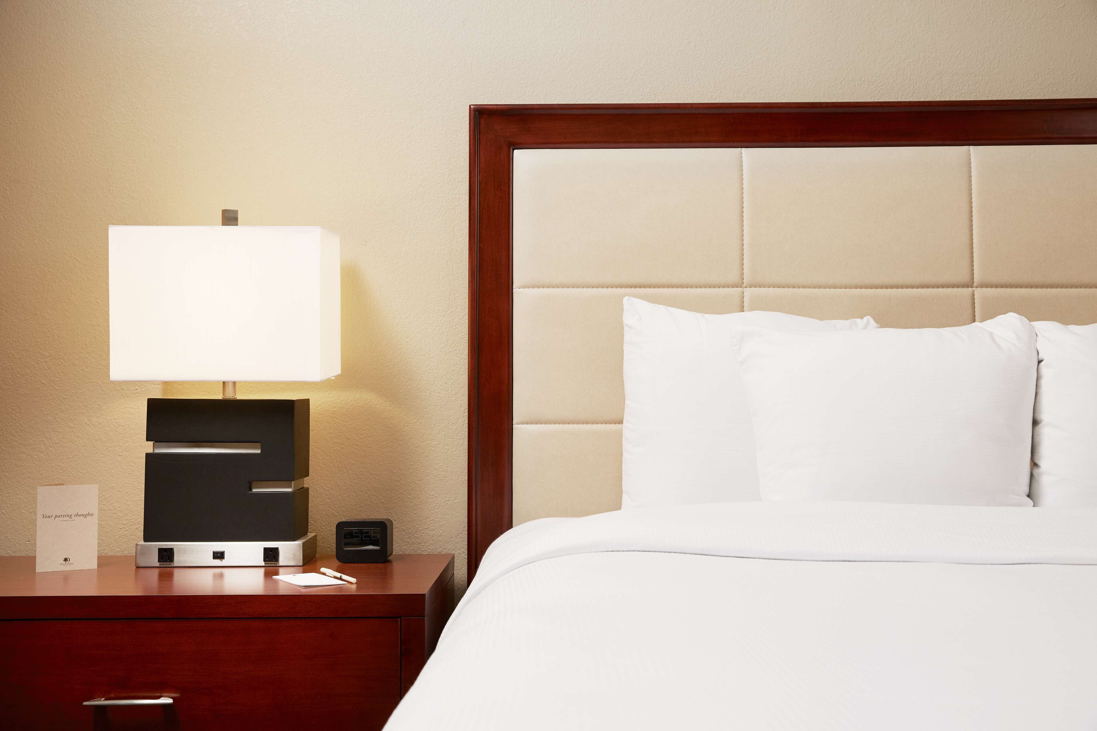 A hotel room with a bed, a bedside table with a lamp, a sign, and a small electronic device.