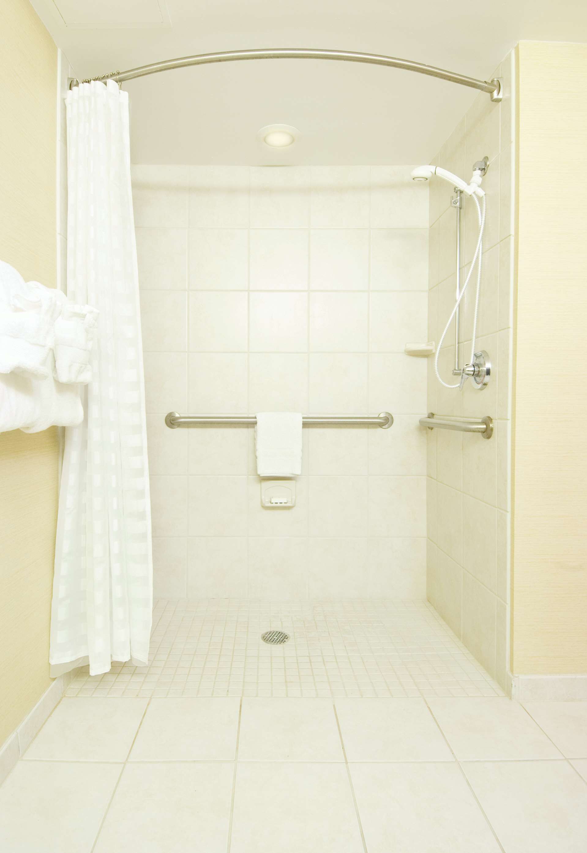 Une douche à l’italienne dotée d’un pommeau de douche incurvé, de barres d’appui et d’un sol carrelé de carreaux blancs.
