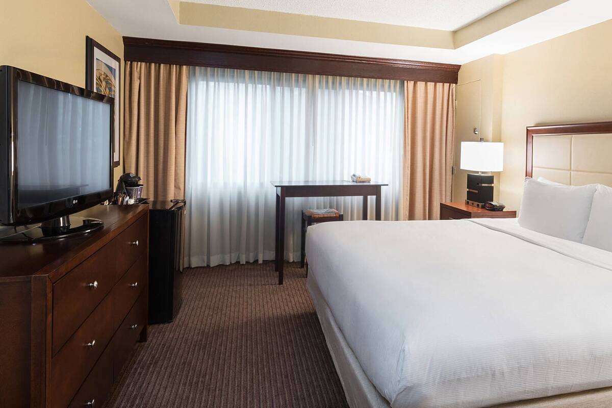 A hotel room with a large bed, a television on a wooden dresser, a desk with a lamp, and a window with curtains.