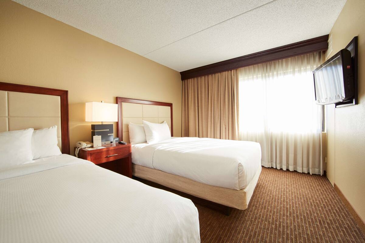 A hotel room with two beds, a bedside table with a lamp, a TV mounted on the wall, and a window with curtains.