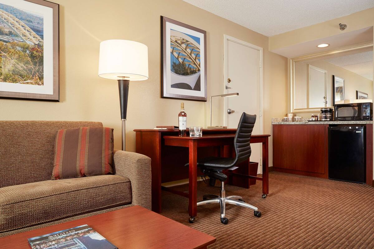 A hotel room with a sofa, a desk, a chair, a lamp, a framed picture, and a kitchenette.