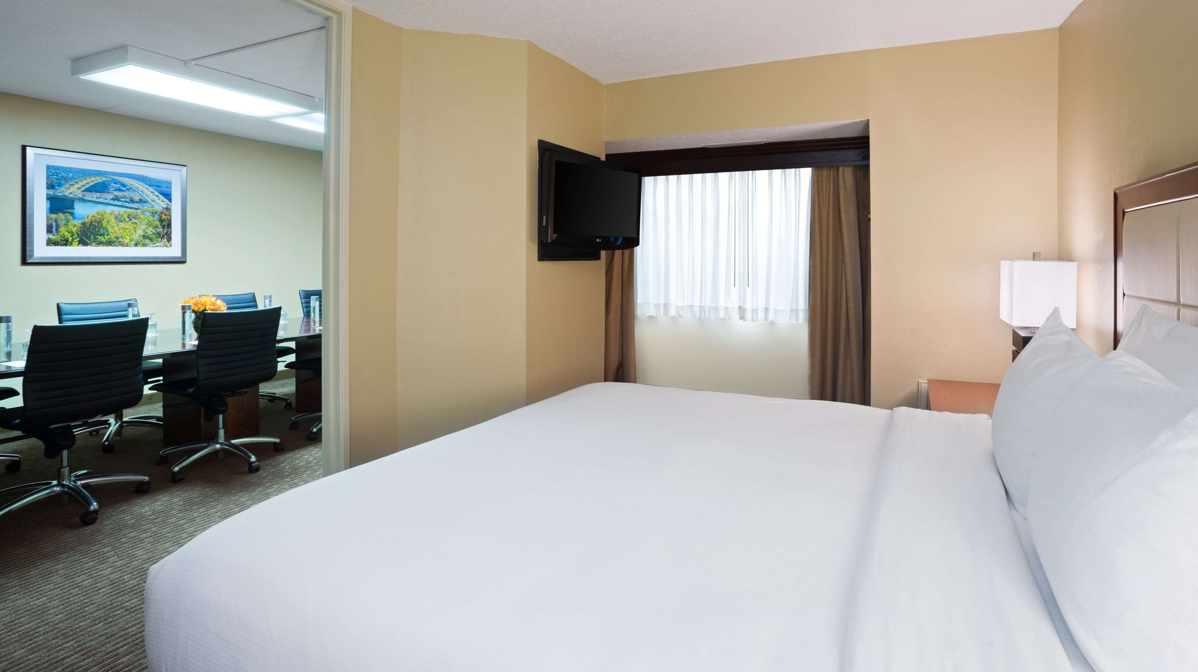 Quarto de hotel com uma cama grande, uma escrivaninha com duas cadeiras, uma TV fixada na parede e uma janela com cortinas.