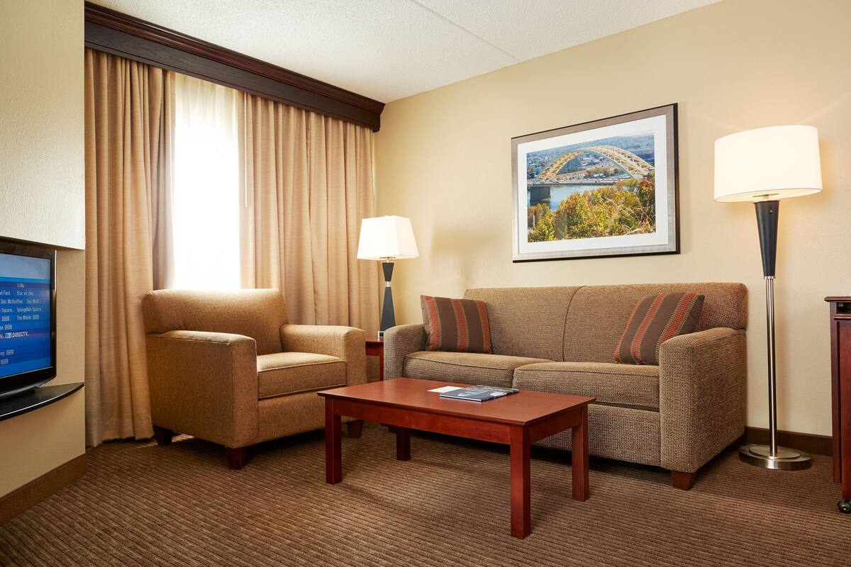 A hotel room with a sofa, armchair, coffee table, TV, and a framed picture on the wall.