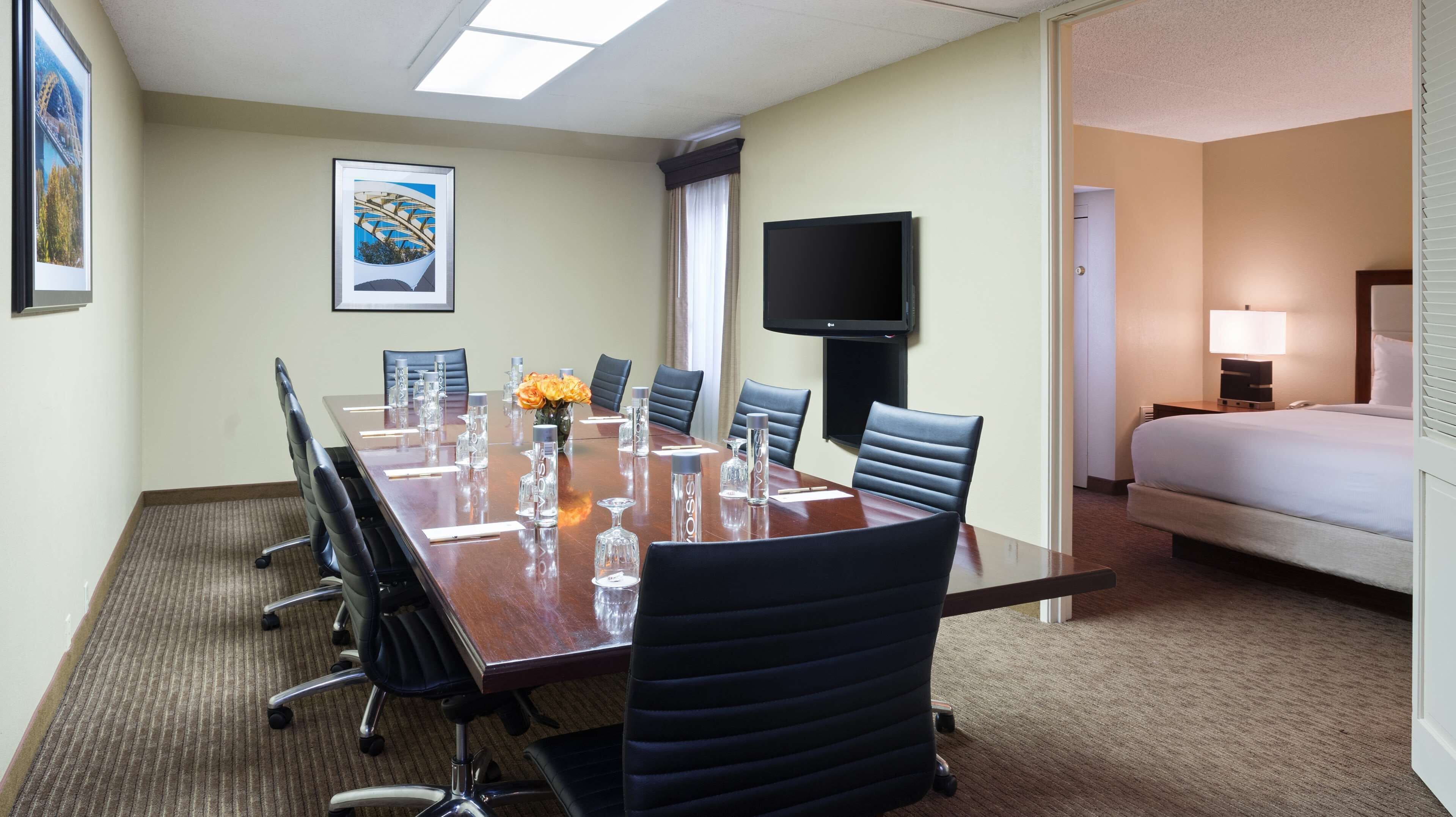 A hotel conference room with a long table, chairs, a TV, and a bed in the adjacent room.