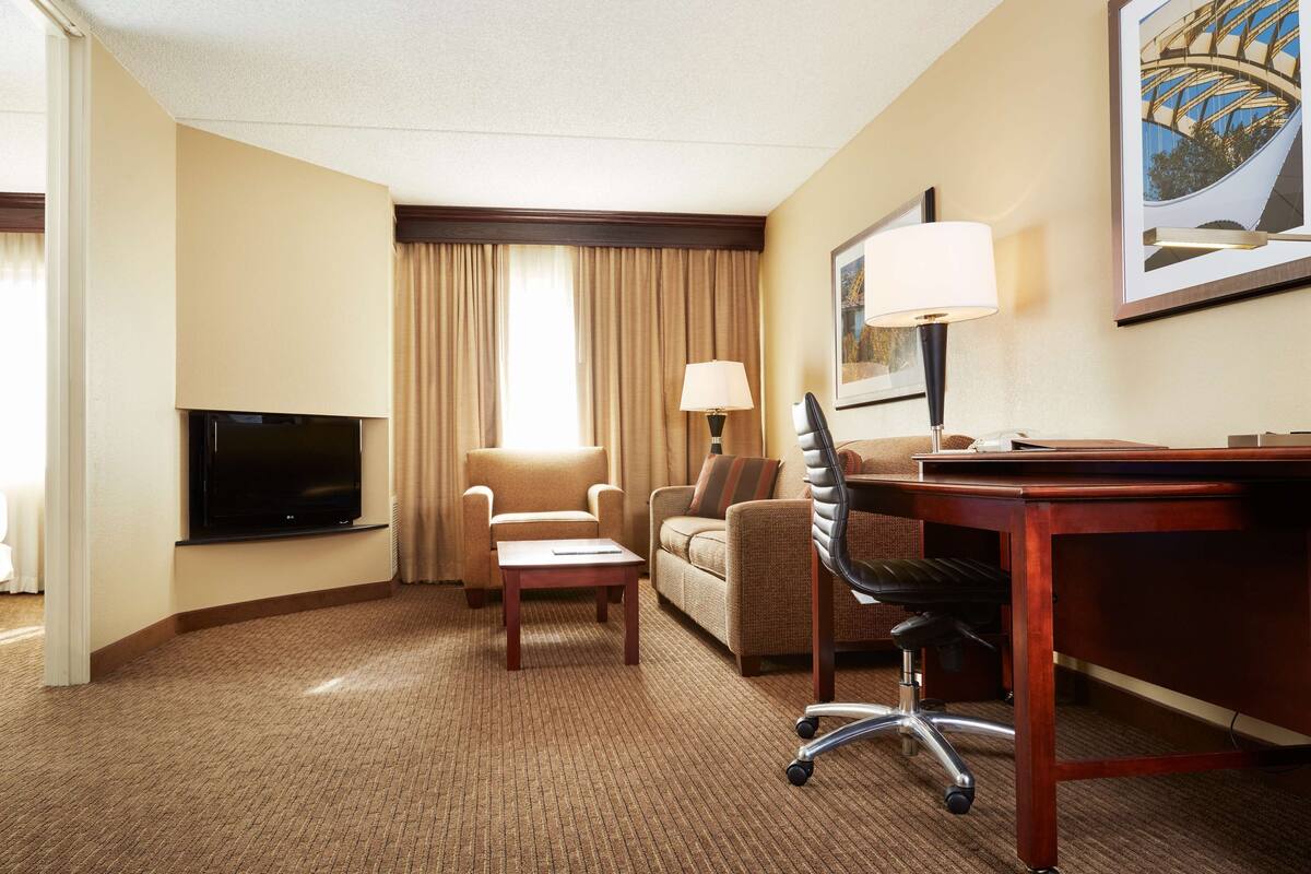 A hotel room with a flat-screen TV, two sofas, a desk with a chair, a lamp, and a painting on the wall.