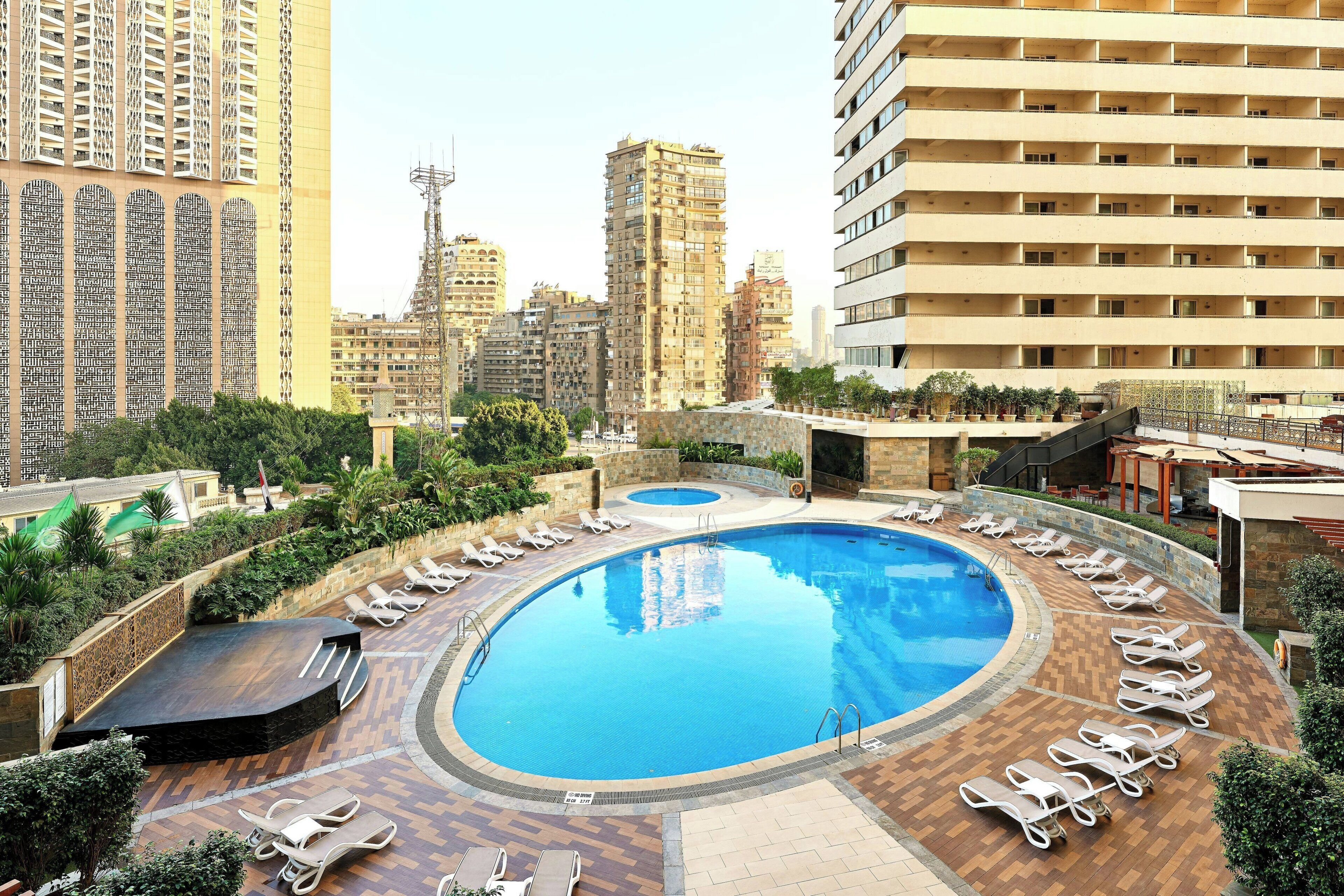 A rooftop swimming pool with sun loungers, surrounded by greenery and high-rise buildings.