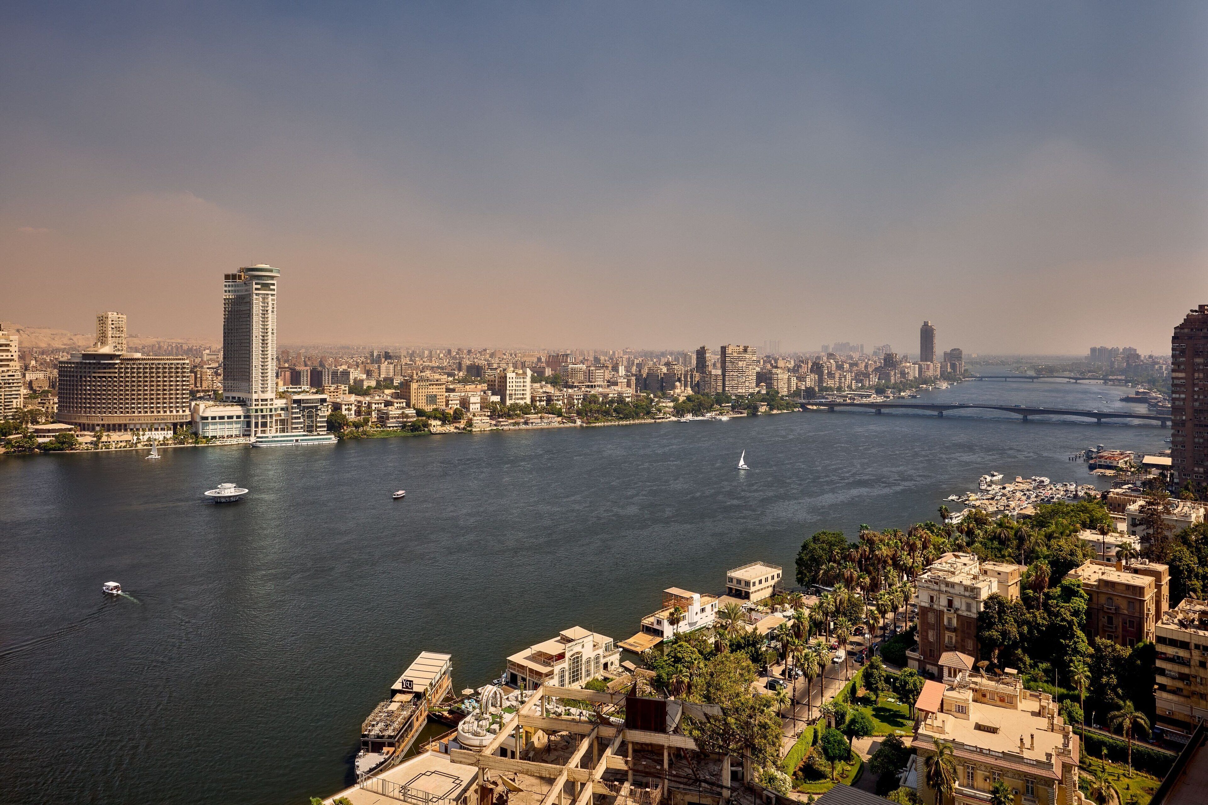 A river with boats, a bridge, and a cityscape with buildings and palm trees.