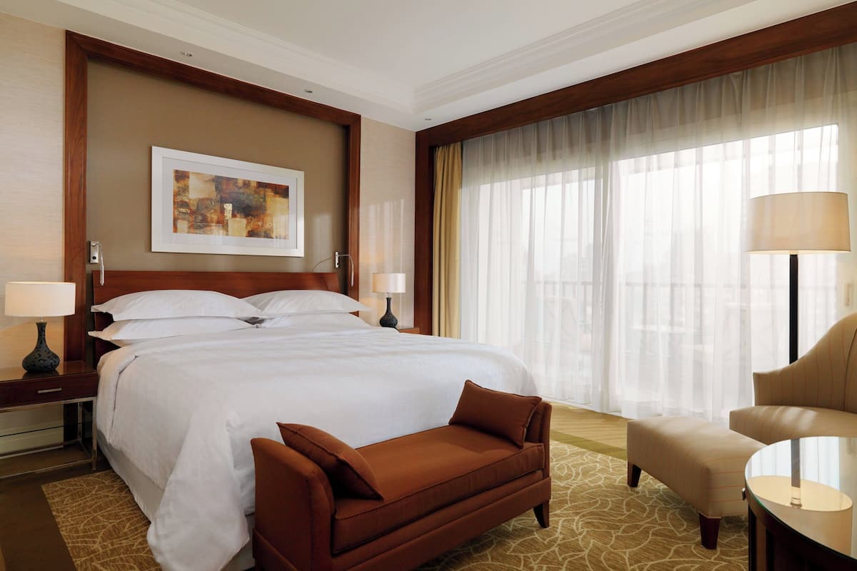Habitación de hotel con una cama grande, un sofá marrón, una mesa redonda y un ventanal con cortinas translúcidas.